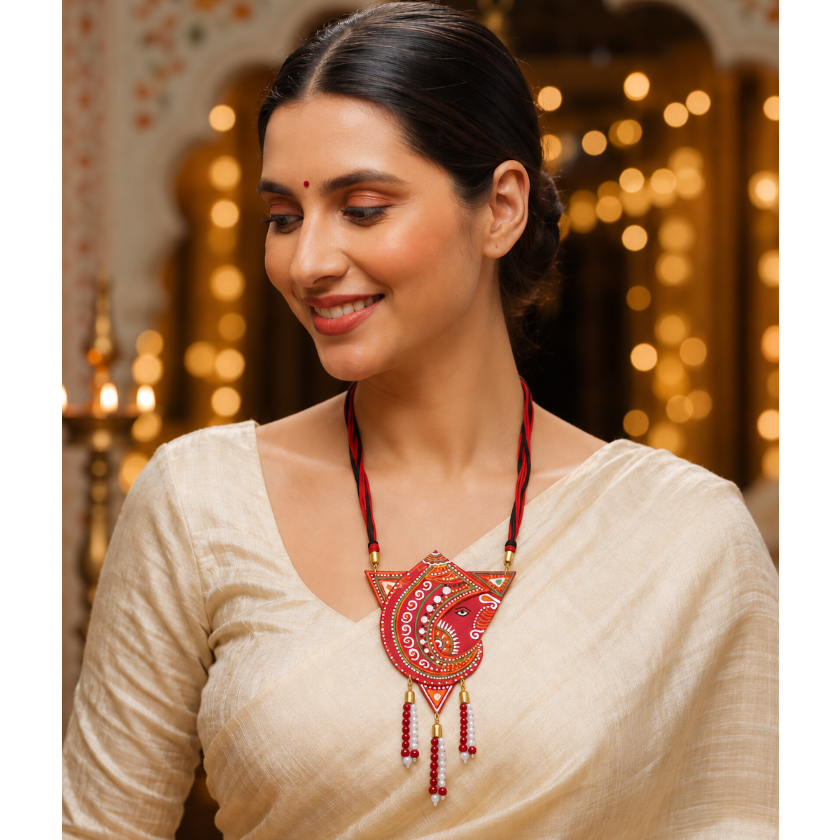 A woman wearing a cream-colored traditional outfit and a prominent red and gold embroidered necklace with dangling red beads, posed in a warmly lit indoor setting with blurred golden lights in the background.